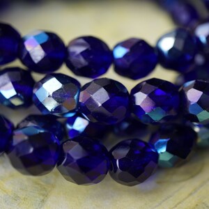 Puede incluir: Una hebra de cuentas de vidrio facetadas de color azul oscuro con brillo iridiscente. Las cuentas son redondas y tienen una forma ligeramente irregular.