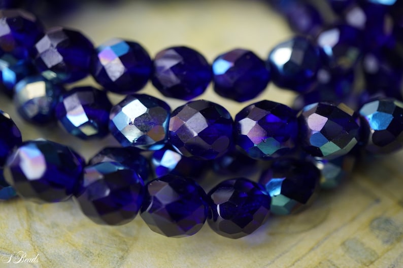 Puede incluir: Una hebra de cuentas de vidrio facetadas de color azul oscuro con brillo iridiscente. Las cuentas son redondas y tienen una forma ligeramente irregular.