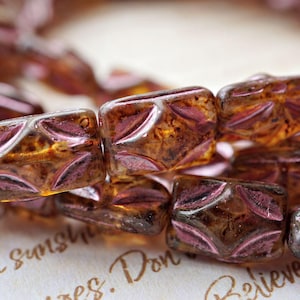 May include: Close-up of a strand of square beads. The beads are a translucent amber color with a pinkish-purple floral design on each face. The beads are strung together, creating a decorative element.