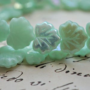 May include: Close-up of a strand of light green leaf-shaped beads. The beads have a subtle iridescent sheen and are arranged in a row. The background is a vintage paper with cursive writing.