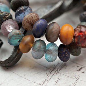 Peut inclure: Gros plan d'un collier composé de perles de verre de différentes couleurs. Les perles sont rondes et ont une finition marbrée et irisée. Le collier est enfilé sur une chaîne en métal.