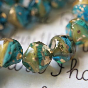 May include: Close-up of a strand of faceted beads. The beads are a mix of teal, gold, and olive green, with a speckled, iridescent finish. The beads are strung together, suggesting jewelry or craft supplies.