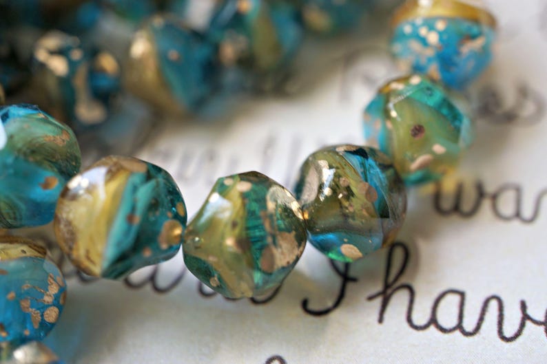May include: Close-up of a strand of faceted beads. The beads are a mix of teal, gold, and olive green, with a speckled, iridescent finish. The beads are strung together, suggesting jewelry or craft supplies.