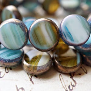 May include: Close-up of a collection of round, flat beads. The beads have a marbled appearance with shades of blue, green, and white. Each bead is framed with a bronze-colored metal. The beads are likely for jewelry making.