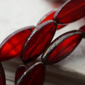 May include: Close-up of a strand of red glass beads. Each bead is an elongated oval shape with a metallic silver-coloured edge. The beads are arranged diagonally across a white surface.