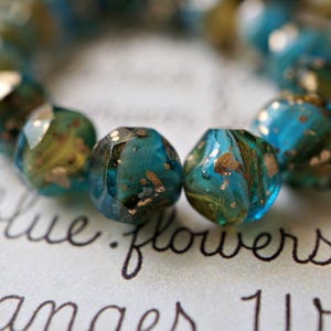 May include: Close-up of a strand of faceted beads. The beads are a mix of teal and gold, with a speckled, marbled appearance. The beads are arranged on a surface with handwritten text, including the words "blue flowers."