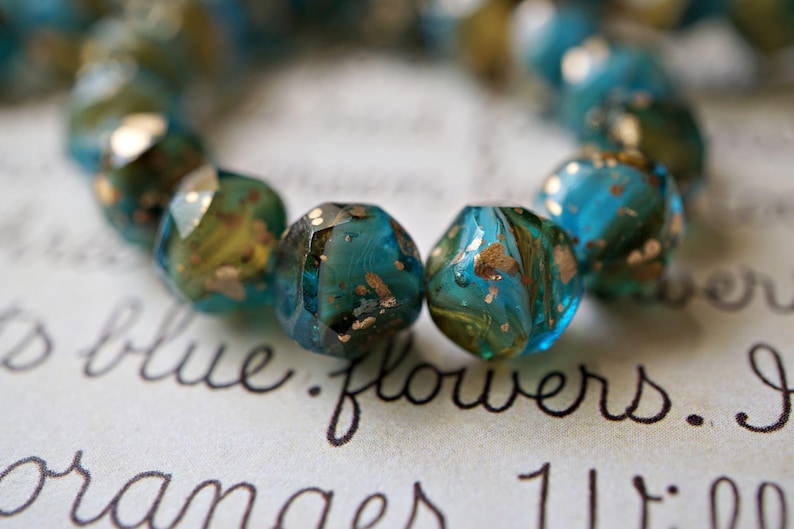 May include: Close-up of a strand of faceted beads. The beads are a mix of teal and gold, with a speckled, marbled appearance. The beads are arranged on a surface with handwritten text, including the words "blue flowers."