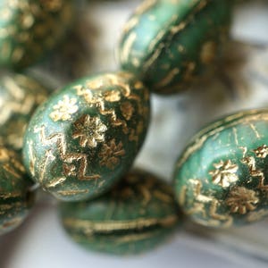 Op de afbeelding: Close-up van verschillende groene kralen met gouden bloempatronen. De kralen hebben een ovale vorm en een licht getextureerd oppervlak. De gouden details voegen een vleugje elegantie toe. Geschikt voor het maken van sieraden.