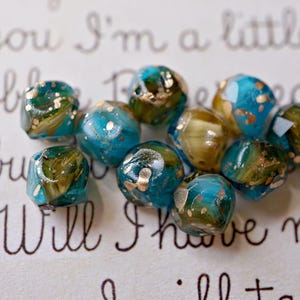May include: A collection of artisan beads in shades of teal, olive green, and gold. The beads have a faceted shape and a marbled appearance, with gold flecks throughout. The background features handwritten script.