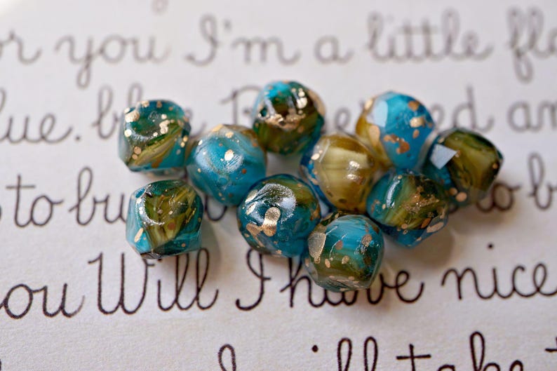 May include: A collection of artisan beads in shades of teal, olive green, and gold. The beads have a faceted shape and a marbled appearance, with gold flecks throughout. The background features handwritten script.