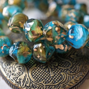 May include: Close-up of a beaded bracelet with faceted, turquoise and olive green beads. The beads have gold flecks and are resting on a decorative, bronze-colored metal heart pendant. Jewelry.