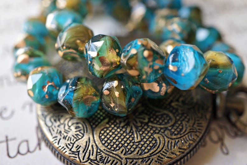 May include: Close-up of a beaded bracelet with faceted, turquoise and olive green beads. The beads have gold flecks and are resting on a decorative, bronze-colored metal heart pendant. Jewelry.