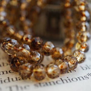 May include: A strand of brown faceted glass beads with a mottled, cloudy appearance. The beads are strung together on a clear thread and are arranged in a loose, flowing pattern.