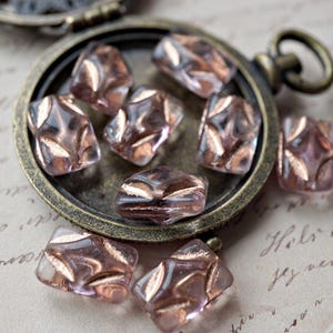 Könnte beinhalten: Eine Sammlung kleiner, irisierender Glasperlen in einem antiken Messingmedaillon. Die Perlen sind hellrosa mit einem dezenten Schimmer und einem zarten Muster.