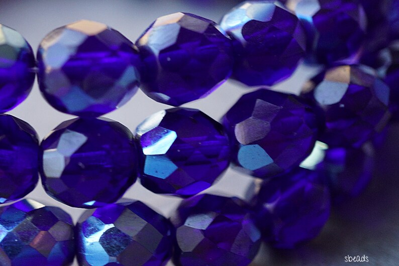 Puede incluir: Primer plano de una hebra de cuentas de vidrio facetadas de color azul iridiscente. Las cuentas son redondas y tienen una superficie brillante y reflectante.