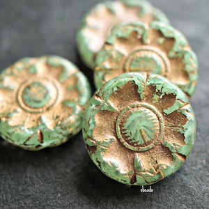 May include: Close-up of several round, antique-style beads with a floral design. The beads have a distressed, aged appearance with a turquoise-green and gold colour scheme. The beads are arranged on a dark grey surface.