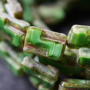 May include: A close-up of a strand of green glass beads with a textured, aged look. The beads are square shaped with a slight curve and have a mottled green color.