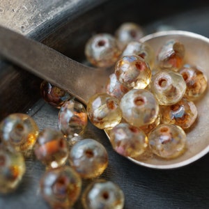 Könnte beinhalten: Nahaufnahme einer Sammlung facettierter, transluzenter Perlen in den Farbtönen Bernstein, Braun und Gold. Die Perlen befinden sich in einem Silberlöffel, wobei einige herausfallen. Die Perlen haben ein Loch in der Mitte und sind wahrscheinlich für die Schmuckherstellung.