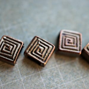 May include: Four square-shaped metal beads with a copper finish and a silver-colored geometric design. The beads have a raised, intricate pattern that resembles a maze or spiral.