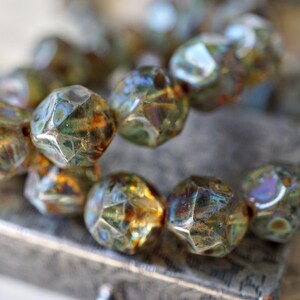 May include: Close-up of a strand of faceted beads. The beads have a mix of colors, including green, gold, and hints of blue. The beads are resting on a textured surface, suggesting a jewelry or craft item.