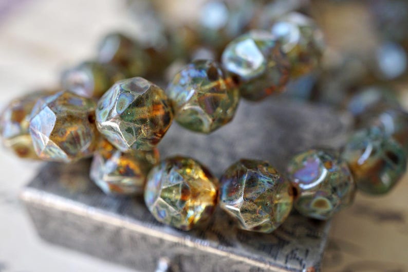 May include: Close-up of a strand of faceted beads. The beads have a mix of colors, including green, gold, and hints of blue. The beads are resting on a textured surface, suggesting a jewelry or craft item.