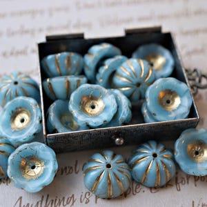 May include: A collection of light blue flower-shaped beads with gold accents, some inside a small metal box. The beads have a textured, ribbed design and a central hole. These decorative beads are ideal for jewelry making and craft projects.