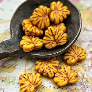 May include: A collection of yellow and brown glass leaf-shaped beads in a metal spoon. The beads have a textured surface and are arranged in a pile.