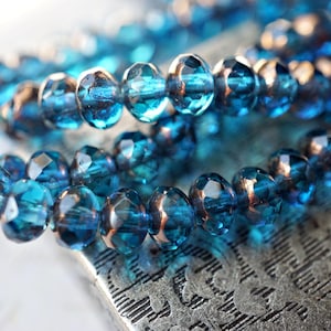 May include: Close-up of a strand of faceted, teal-colored beads with copper-colored accents. The beads are strung together, creating a necklace or bracelet. The beads are resting on a textured, silver surface.