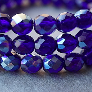 Puede incluir: Primer plano de una hebra de cuentas de cristal facetadas de color azul iridiscente. Las cuentas son redondas y tienen un acabado brillante.