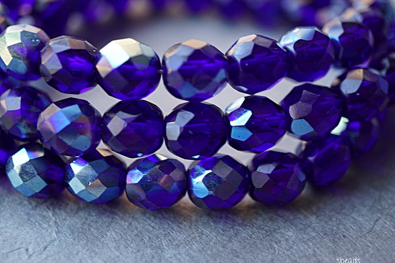 Puede incluir: Primer plano de una hebra de cuentas de cristal facetadas de color azul iridiscente. Las cuentas son redondas y tienen un acabado brillante.