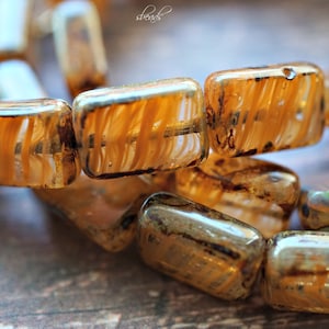 May include: A close-up of a strand of amber-colored glass beads with a striated pattern. The beads are rectangular and have a translucent appearance.