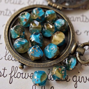 May include: A collection of turquoise and gold-flecked glass beads displayed in a vintage bronze-colored locket. The beads have a marbled appearance and are scattered on a paper background with handwritten script.