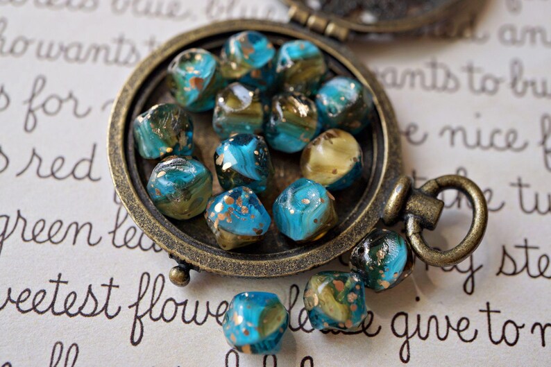 May include: A collection of turquoise and gold-flecked glass beads displayed in a vintage bronze-colored locket. The beads have a marbled appearance and are scattered on a paper background with handwritten script.