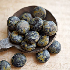 May include: A close-up of a collection of round, black and gold speckled glass beads in a vintage metal spoon. The beads are scattered on a light brown surface.