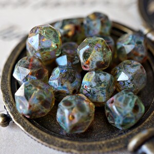 May include: A collection of faceted glass beads in shades of green, blue, and brown, displayed in a vintage bronze-colored locket. The beads have a unique, iridescent quality, reflecting light. The locket is partially open, revealing the beads.