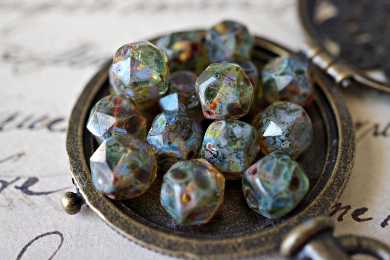 May include: A collection of faceted glass beads in shades of green, blue, and brown, displayed in a vintage bronze-colored locket. The beads have a unique, iridescent quality, reflecting light. The locket is partially open, revealing the beads.