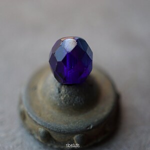 Puede incluir: Una cuenta de vidrio facetada de color morado oscuro con un acabado brillante. La cuenta est&aacute; sentada sobre una peque&ntilde;a base redonda, gris y texturizada.