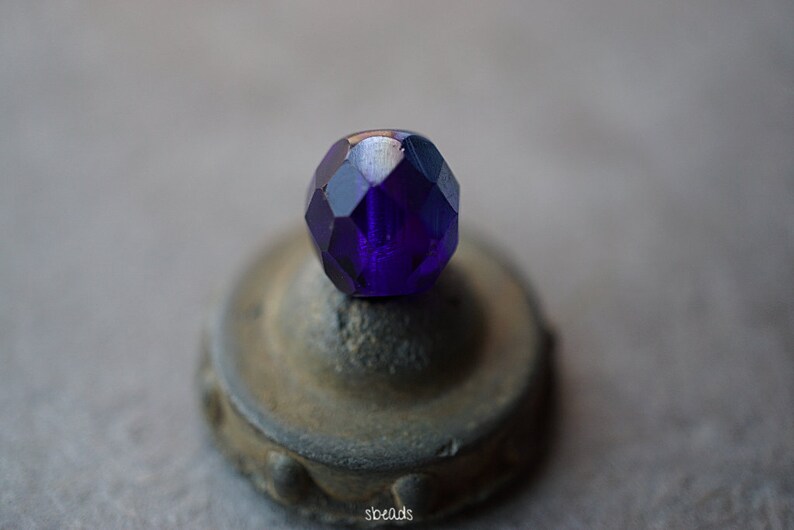 Puede incluir: Una cuenta de vidrio facetada de color morado oscuro con un acabado brillante. La cuenta est&aacute; sentada sobre una peque&ntilde;a base redonda, gris y texturizada.