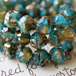 May include: Close-up of a bracelet made of faceted beads. The beads are a mix of teal, gold, and olive green, with a speckled, iridescent finish. The bracelet is resting on a surface with handwritten text.