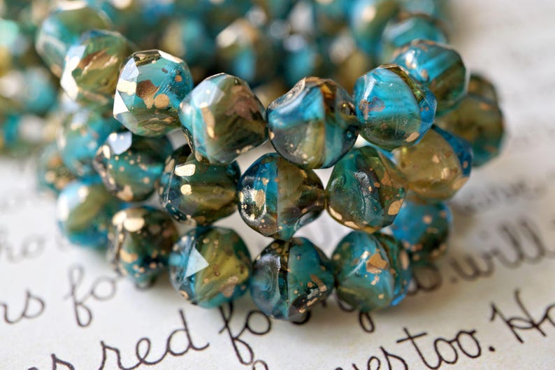 May include: Close-up of a bracelet made of faceted beads. The beads are a mix of teal, gold, and olive green, with a speckled, iridescent finish. The bracelet is resting on a surface with handwritten text.