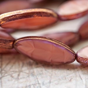 May include: A close-up of a necklace made of pink and copper-toned oval beads. The beads have a metallic sheen and are strung together on a clear thread.