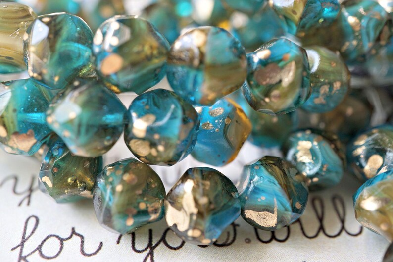 May include: Close-up of a strand of faceted beads. The beads are a mix of teal, blue, and gold. The beads have an irregular shape and are strung together. The background has the words "for you" in cursive.