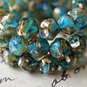 May include: Close-up of a bracelet made of faceted beads. The beads are a mix of teal, blue, and gold, with a speckled, iridescent finish. The beads are arranged in a circular pattern, suggesting a bracelet or necklace.