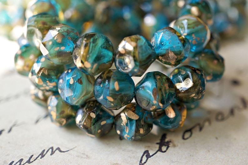 May include: Close-up of a bracelet made of faceted beads. The beads are a mix of teal, blue, and gold, with a speckled, iridescent finish. The beads are arranged in a circular pattern, suggesting a bracelet or necklace.