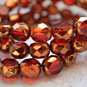 May include: A strand of faceted glass beads in a deep red color with a metallic gold finish. The beads are round and have a faceted design.