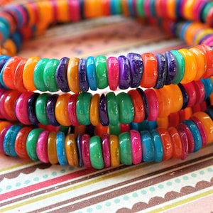 May include: A close-up of a multi-colored strand of round beads. The beads are made of a smooth, shiny material and are arranged in a random pattern of orange, yellow, green, blue, purple, and pink.