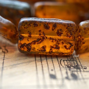 May include: Close-up of a strand of amber beads with a rustic, mottled texture. The beads are a rich, warm brown color and have a translucent quality. The beads are arranged in a line and are resting on a piece of sheet music.