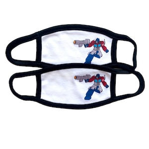 May include: Two white face masks with black elastic ear loops. Each mask features a printed image of Optimus Prime, a red, blue, and gray robot character from the Transformers franchise, holding a gun.