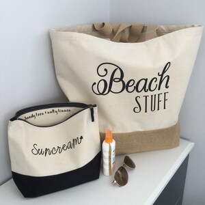 May include: A white canvas tote bag with the text "Beach Stuff" in black cursive lettering. A smaller white canvas zippered pouch with the text "Suncream" in black cursive lettering. The tote bag has a brown burlap bottom. The pouch has a black bottom. A bottle of sunscreen and sunglasses are on a white surface.