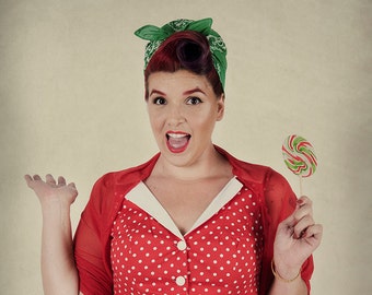 May include: A woman wearing a red polka dot dress with a white collar and a green bandana in her hair. She is holding a colourful lollipop and smiling.
